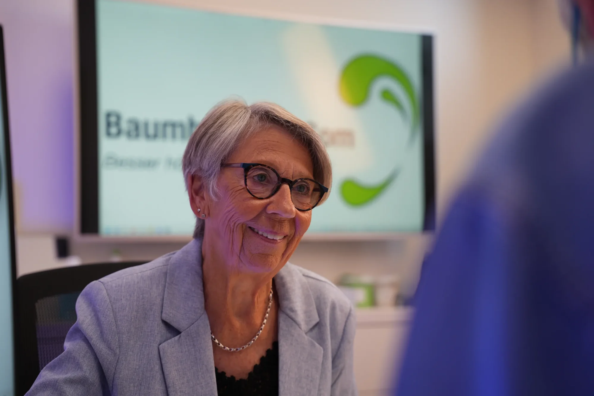Eine Kundin mit Brille lächelt im Beratungsgespräch, hinter ihr ein Bildschirm mit dem Logo von Baumbach HörCom.